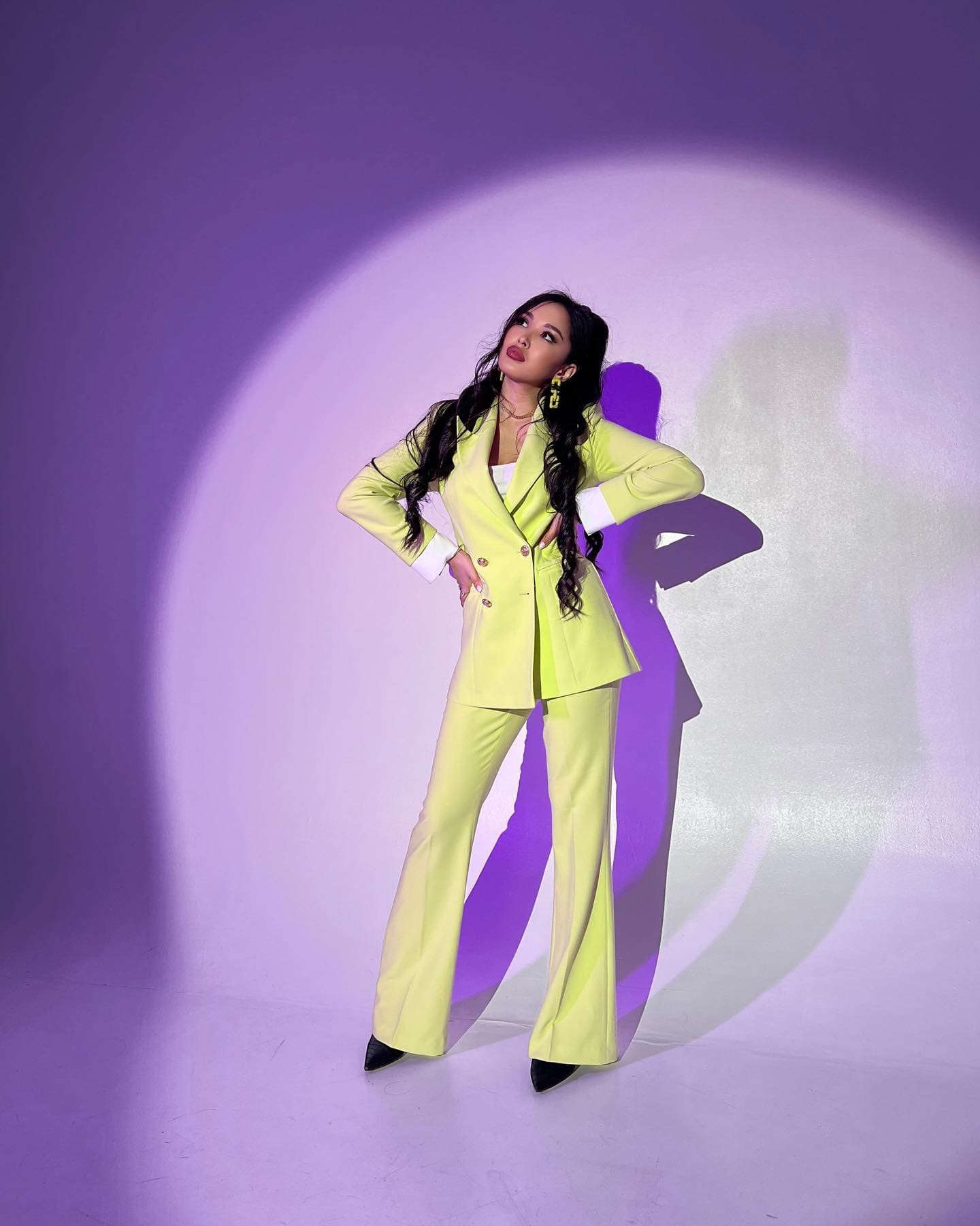 A woman in a yellow tailored suit and black heels poses with hands on hips against a purple-lit backdrop with a circular spotlight.