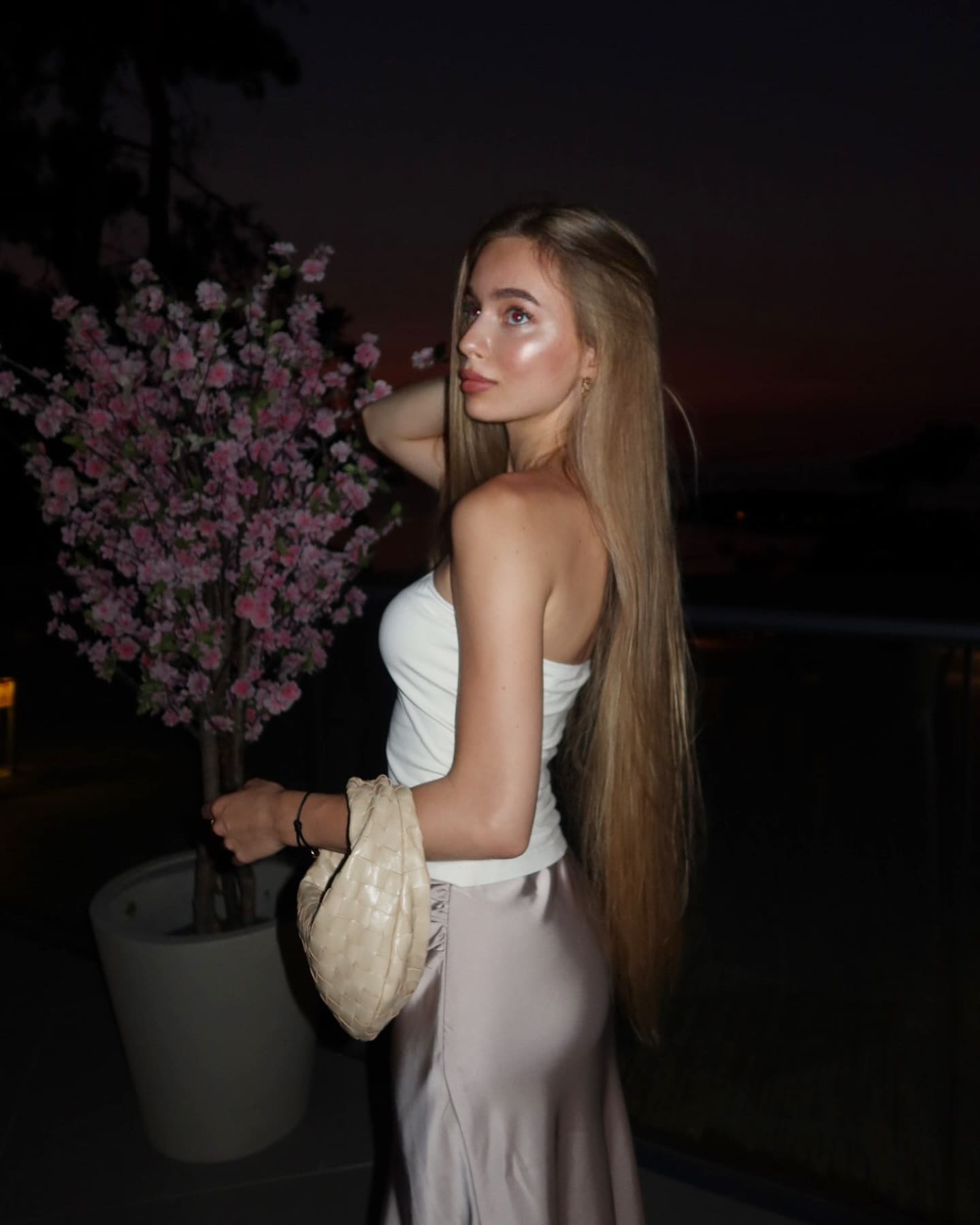 A blonde woman stands in profile at dusk wearing a white strapless top and satin skirt, holding a woven bag and flowering branch.