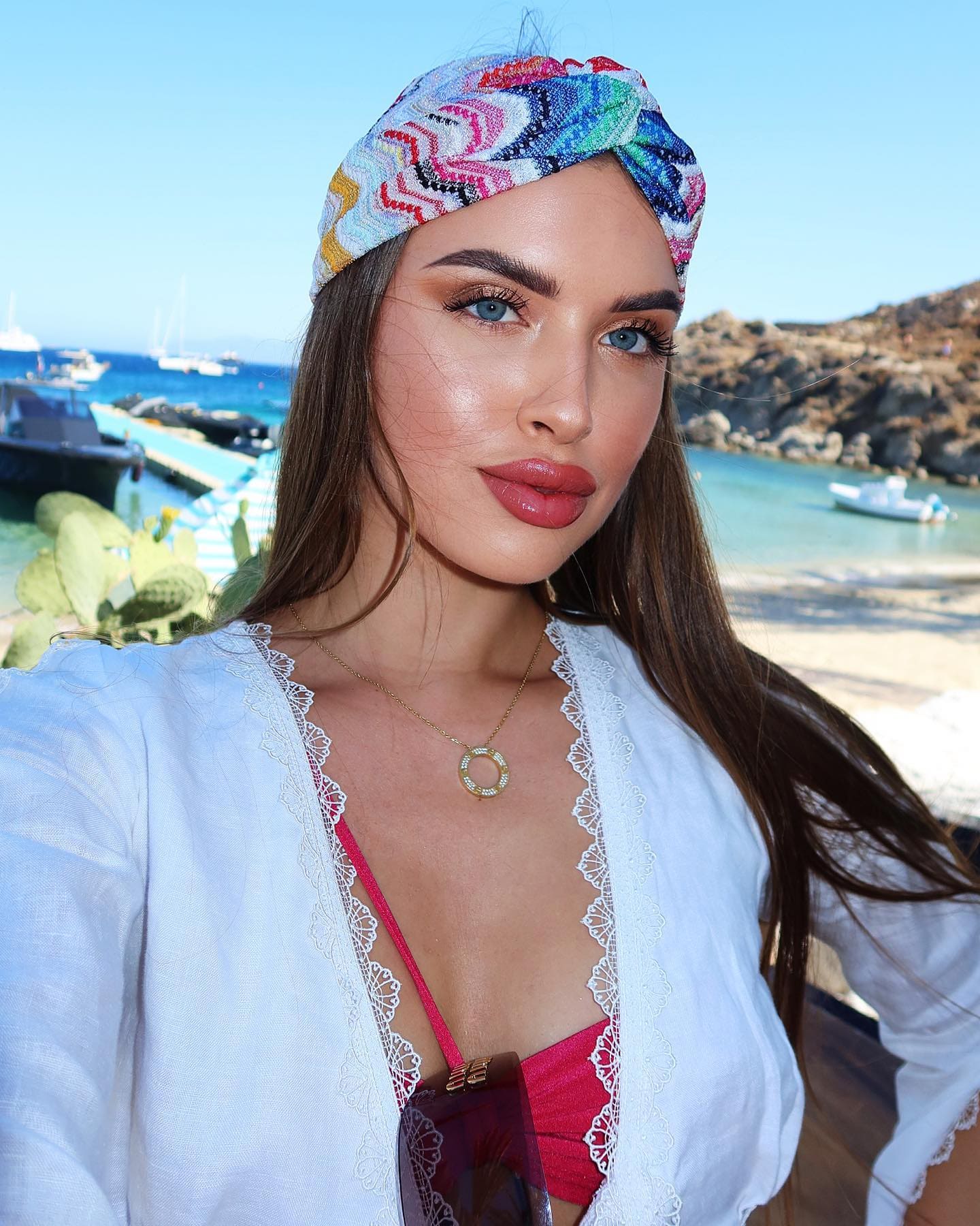 Woman in a white lace cover-up and red bikini with a patterned headscarf poses against a harbor and boats.
