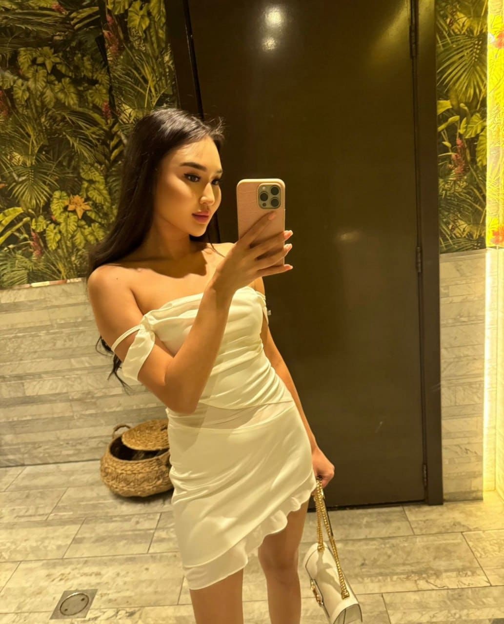 A woman takes a mirror selfie in a white strapless mini dress, holding a phone and small white handbag near patterned wallpaper.