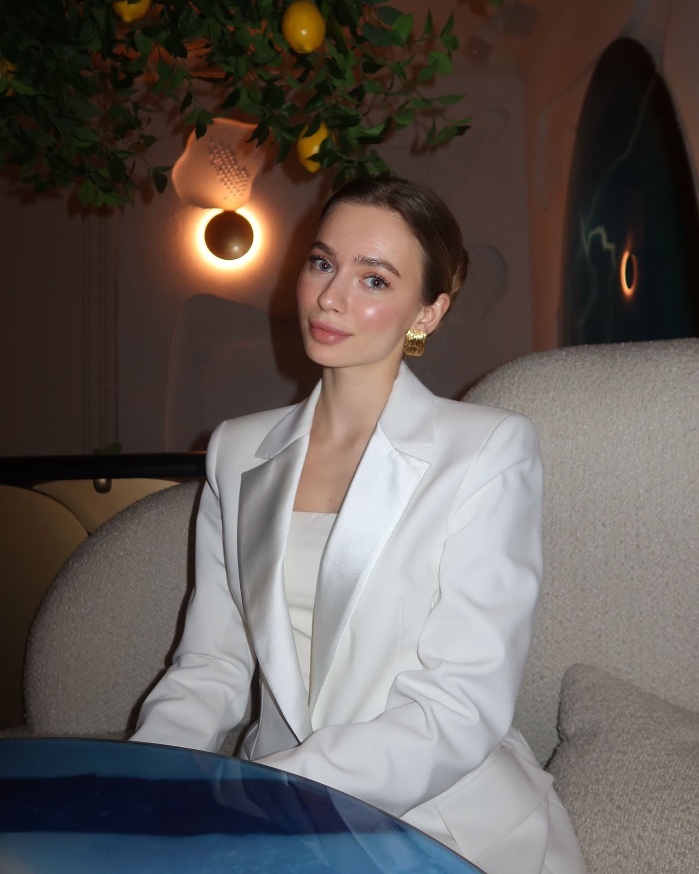 Afira sits on a beige sofa in a white blazer and top, with gold earrings, in a dim room featuring a lemon tree and wall sconce.
