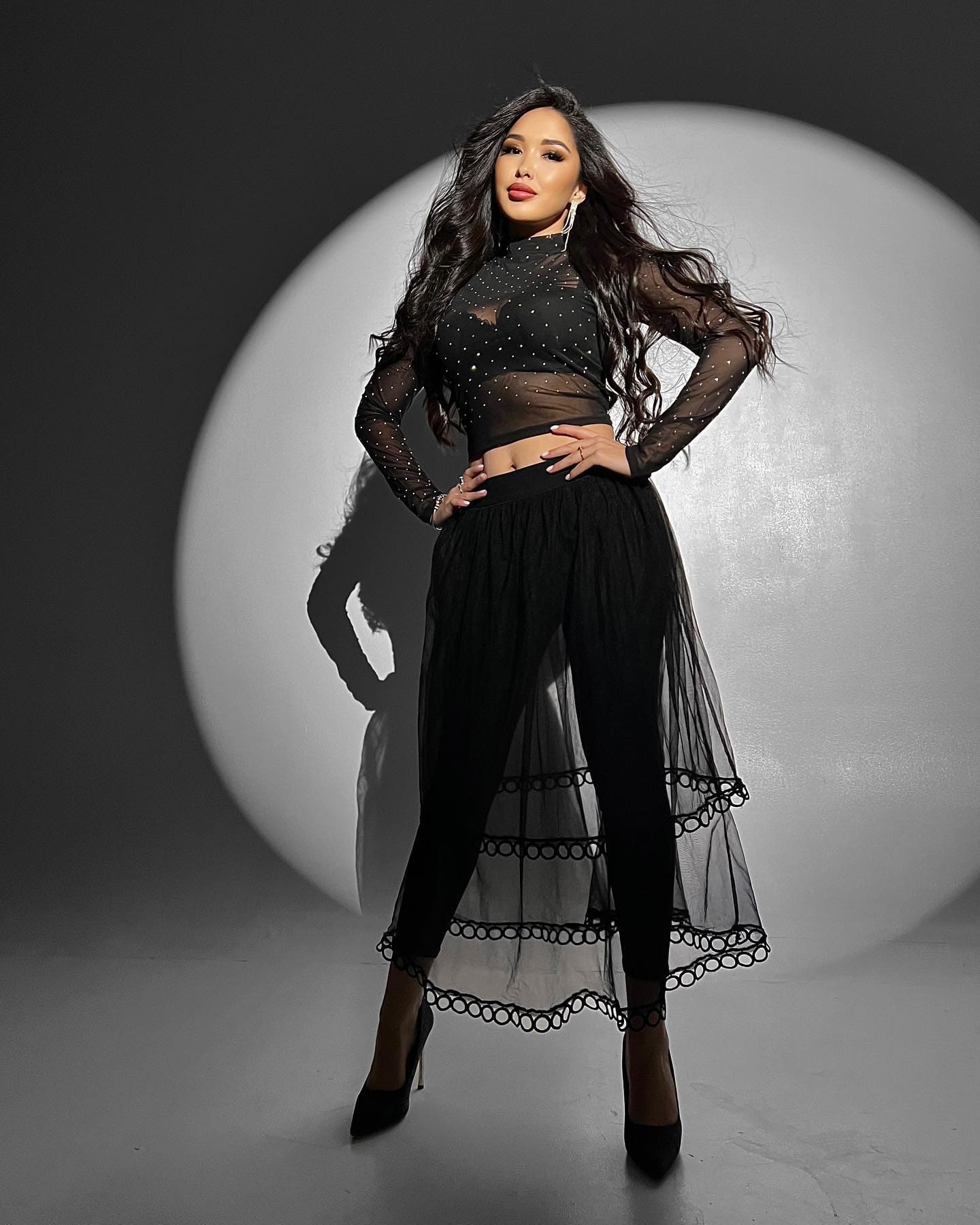 A woman wears a sheer black polka-dot crop top and tulle skirt, posing with hands on hips against a large white circular backdrop.