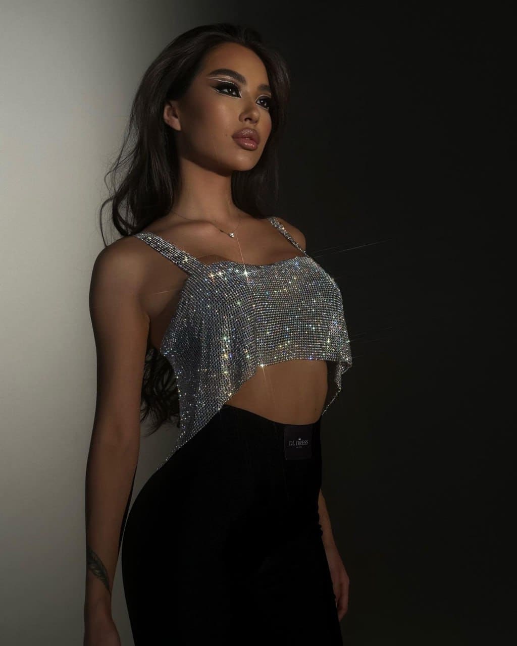 A woman in a silver rhinestone crop top and high-waisted black skirt stands in a studio, showing a forearm tattoo.