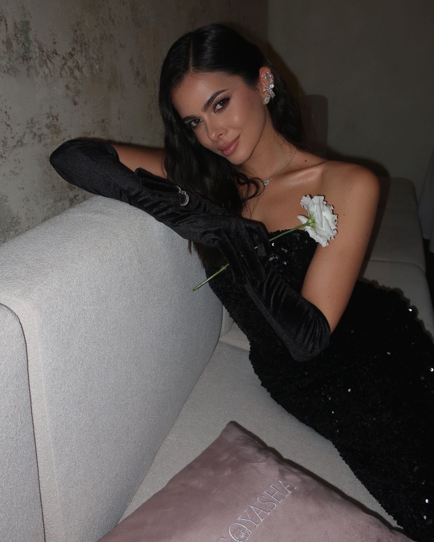 Seated on a light sofa, she wears a strapless black sequined dress and long velvet gloves, holding a white flower near her chest.