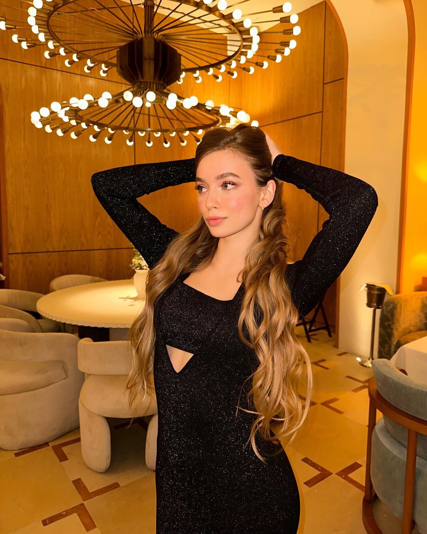 A woman with long wavy brown hair stands indoors in a black sparkly dress with a chest cutout, hands behind her head, near a chandelier.