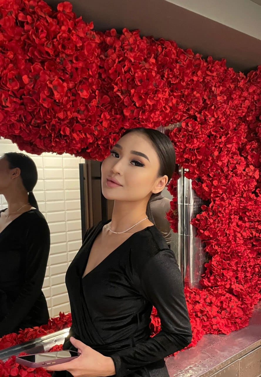 A woman in a black V-neck dress stands before a wall of red flowers, holding a phone with her hair pulled back.