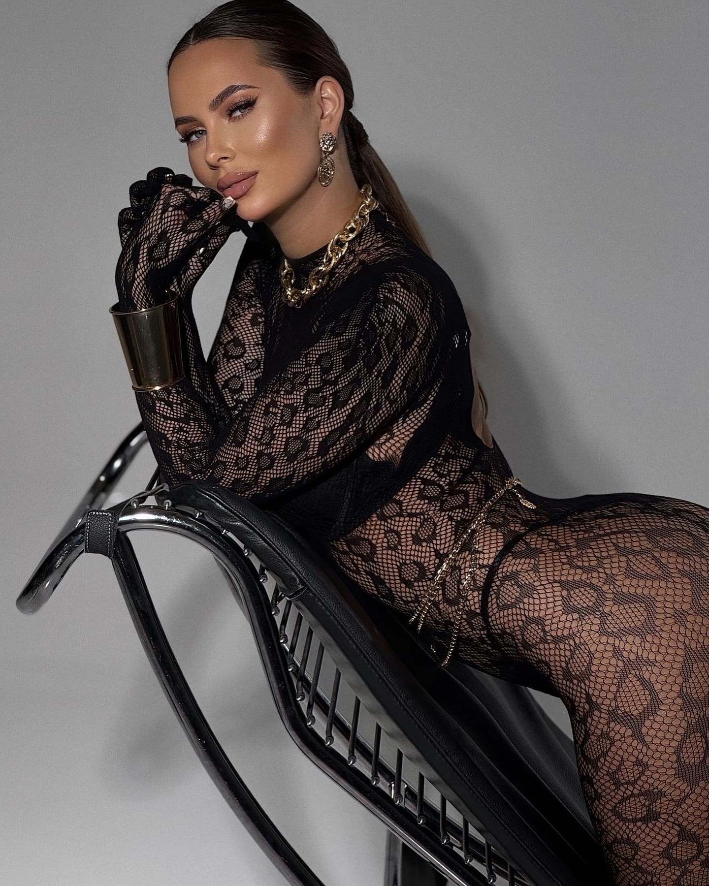 A woman with hair pulled back sits on a black chair in a sheer lace top and tights, wearing gold jewelry and resting a hand near her chin.