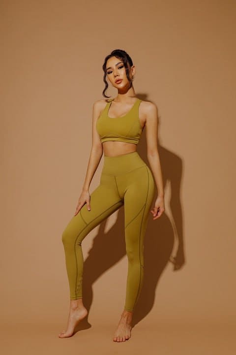 Romma stands barefoot in olive-green activewear, hand on thigh, against a plain tan studio backdrop.