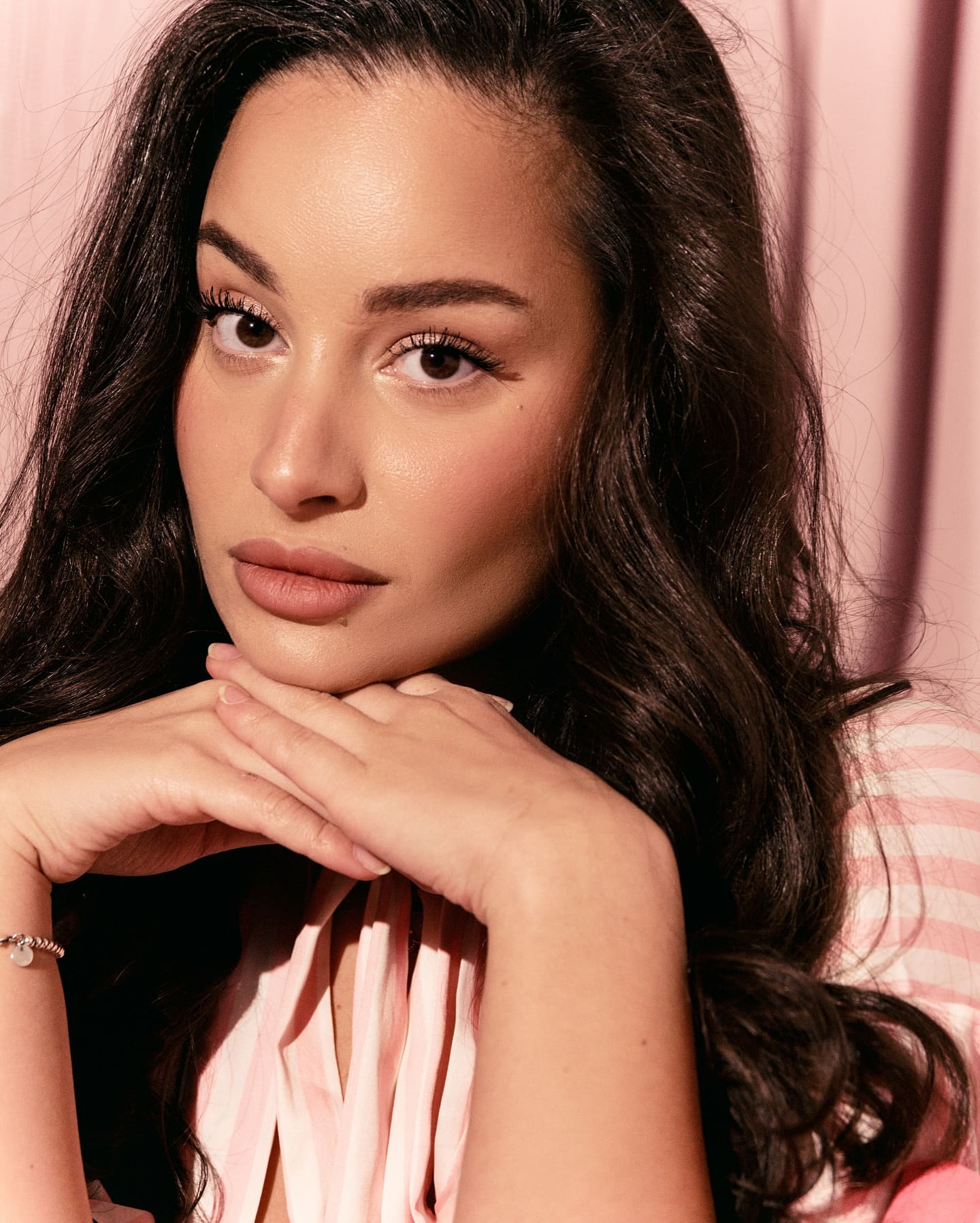 A woman rests her chin on clasped hands while wearing a pink and white striped top against a pink curtain backdrop.