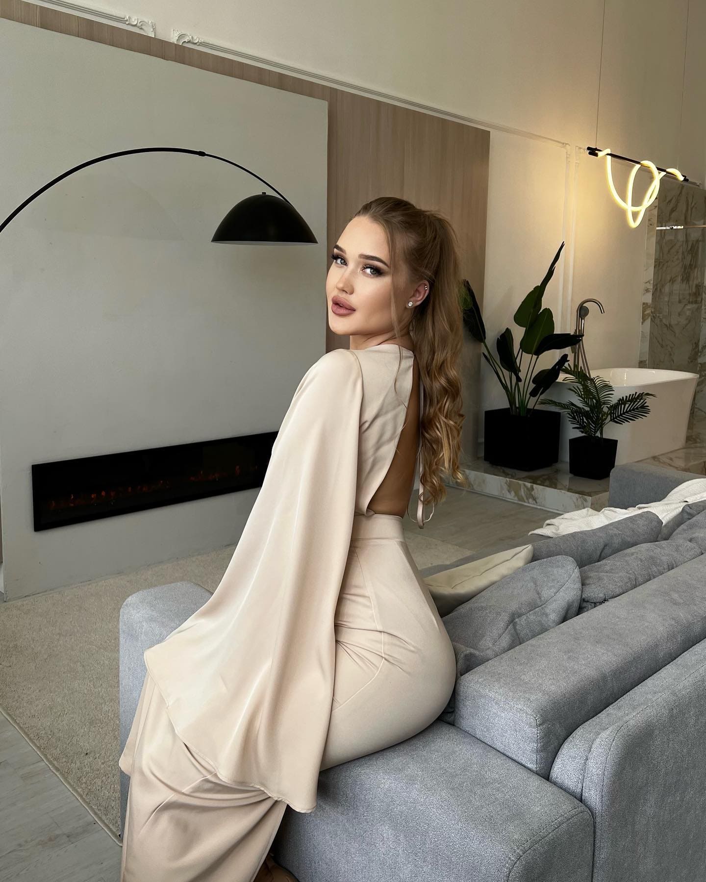 A woman in a beige satin outfit sits on a grey sofa, looking over her shoulder in a modern living room.