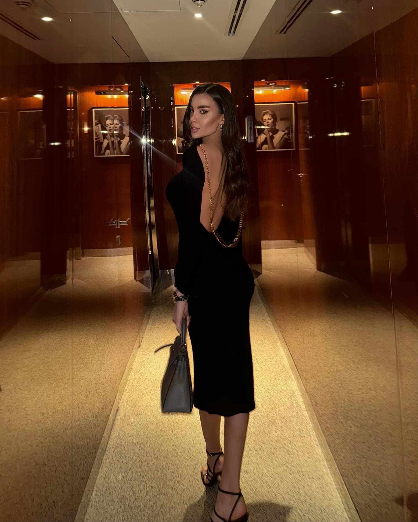 A woman stands in a mirrored hallway wearing a black backless dress with a gold chain, strappy heels, and a gray handbag.