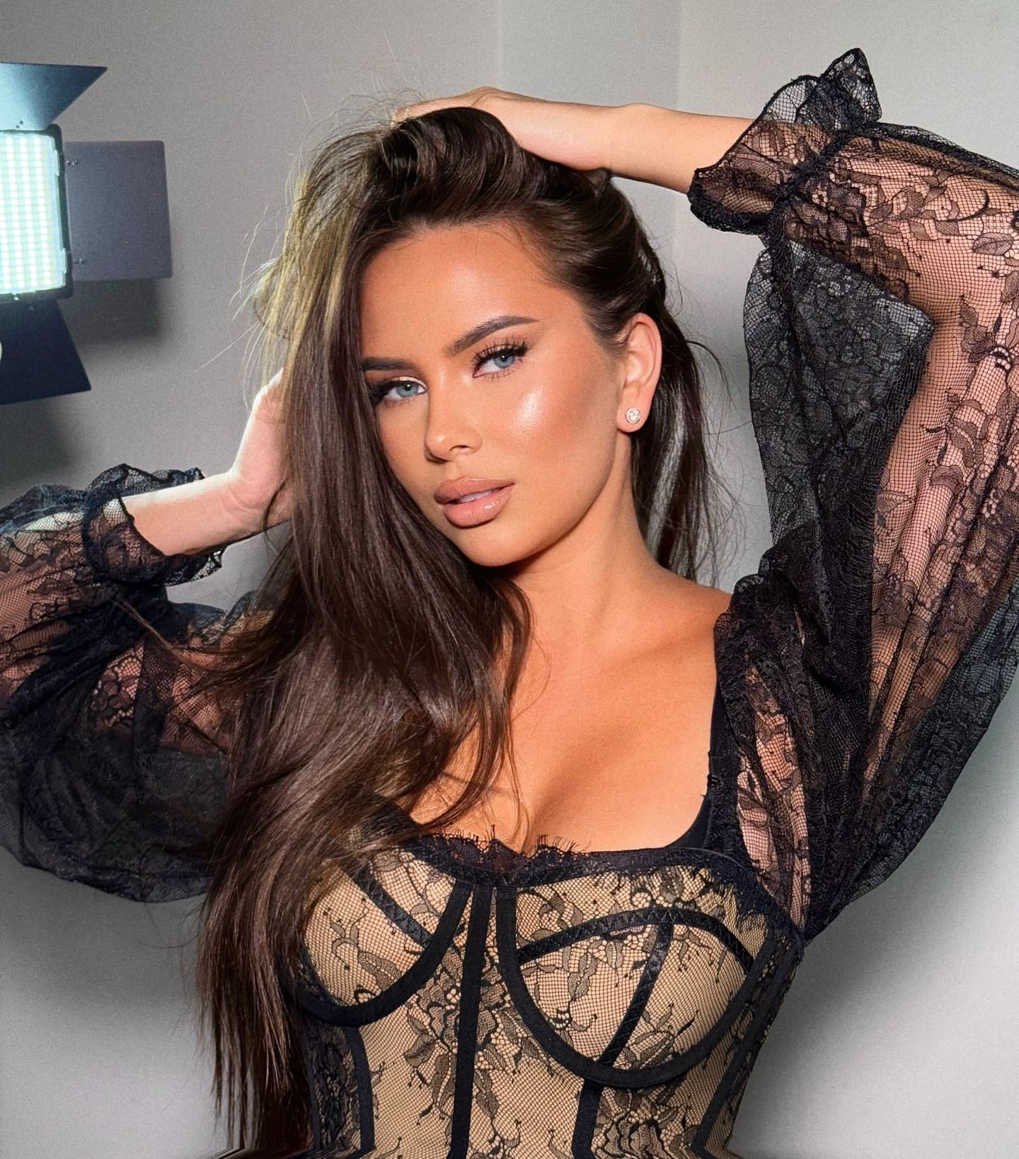 Irenna poses with hands behind her head in a sheer black lace top, standing before a gray wall with a studio light visible to the left.