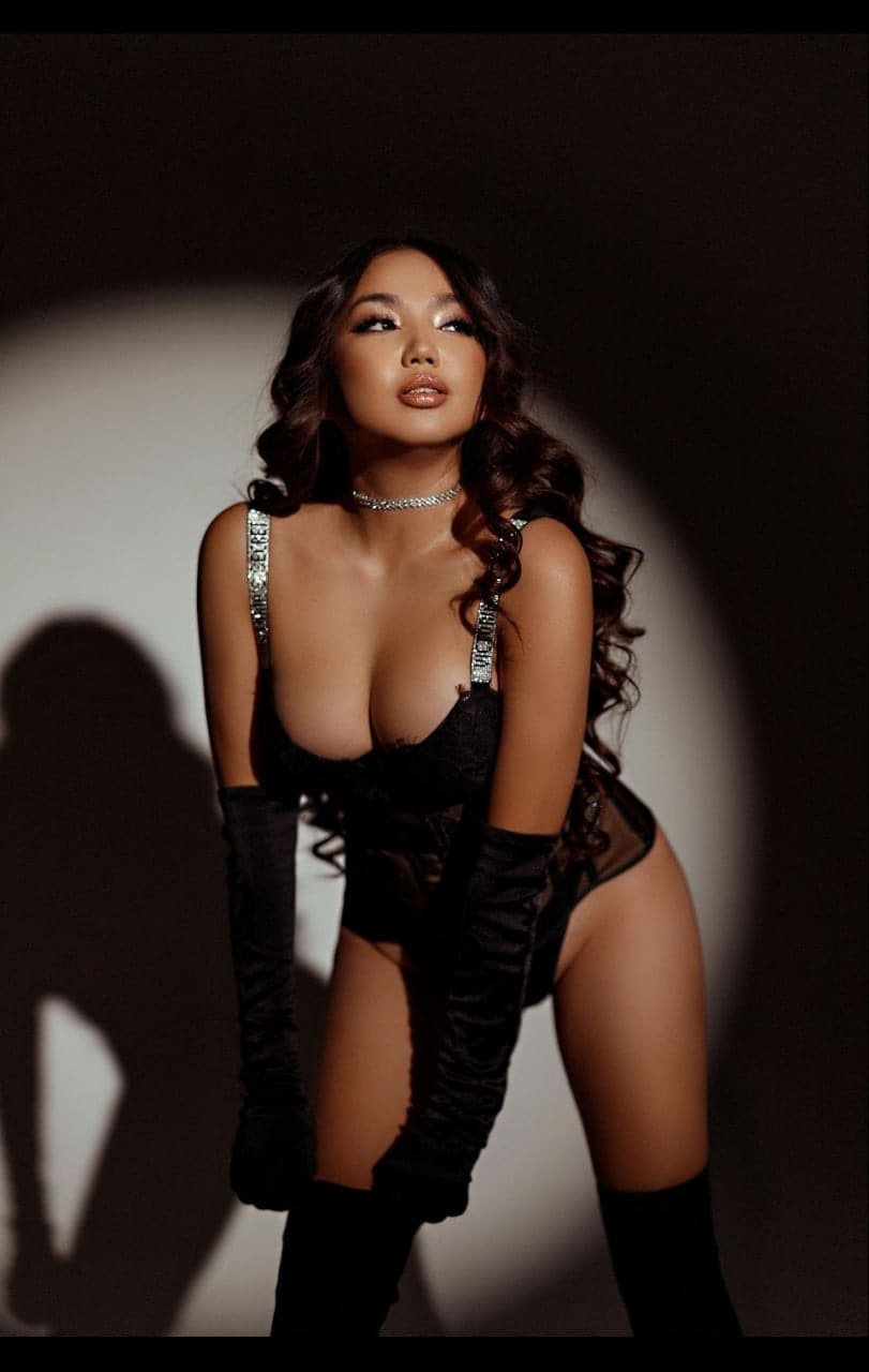 A woman stands in a spotlight wearing a black lace bodysuit, gloves, and stockings, hands resting on her thighs.