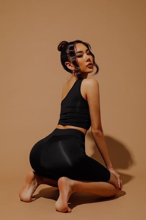 A woman in a black one-shoulder top and leggings kneels barefoot in profile against a beige backdrop.