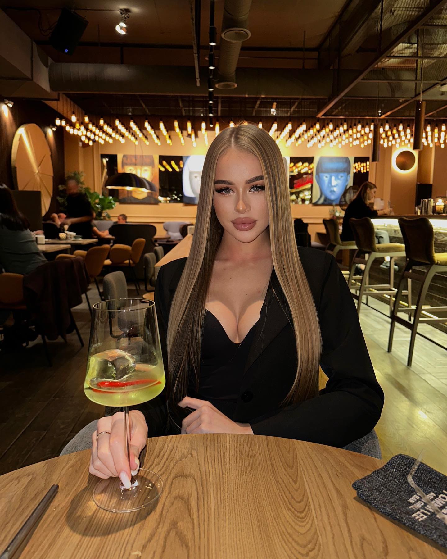 A woman sits at a restaurant table in a black blazer, holding a stemmed glass of yellow drink with a red garnish.