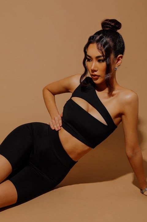 A woman in a black one-shoulder top and shorts sits on a tan floor, one hand on hip, the other supporting her.