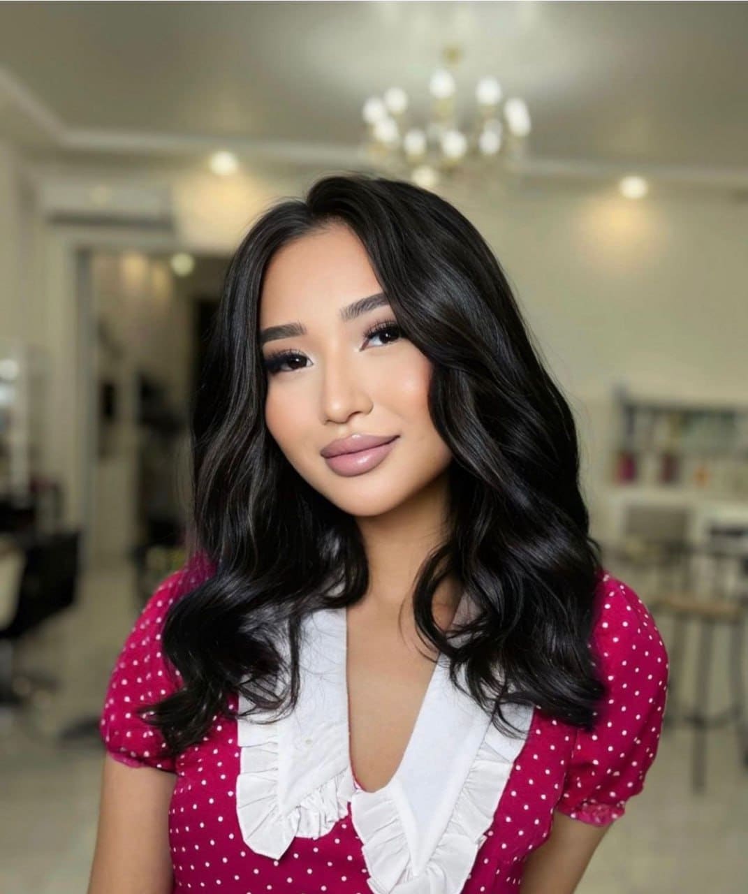 Milana with long dark wavy hair wears a magenta polka-dot dress with a white ruffled collar in a lit indoor space with a chandelier.