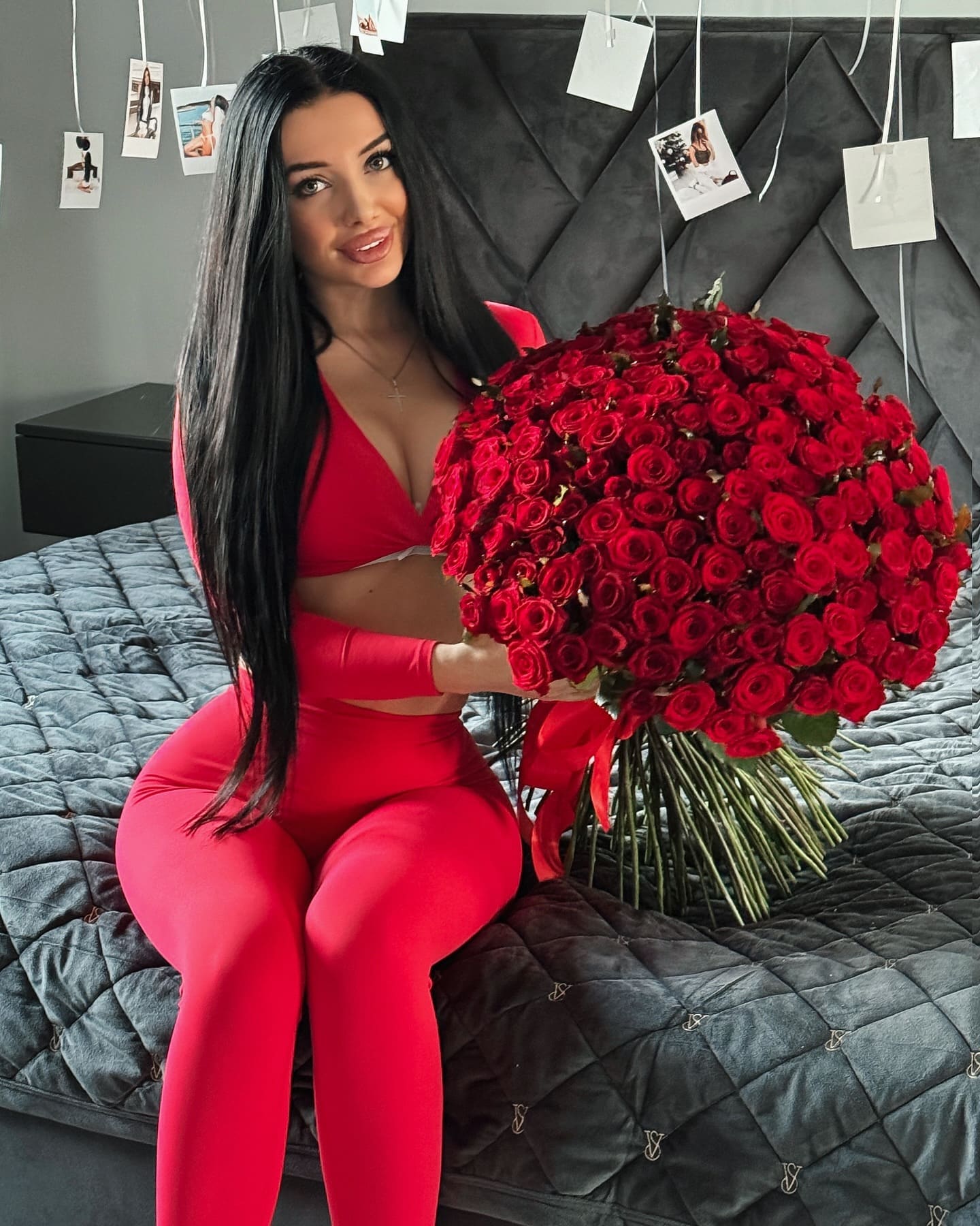 A woman in a red two-piece outfit sits on a grey bed holding red roses, with a dark headboard and photos in the background.