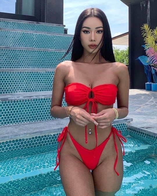 Estell stands in a tiled pool in a red tie-front bikini, hands clasped at her waist, with mosaic steps and plants behind her.