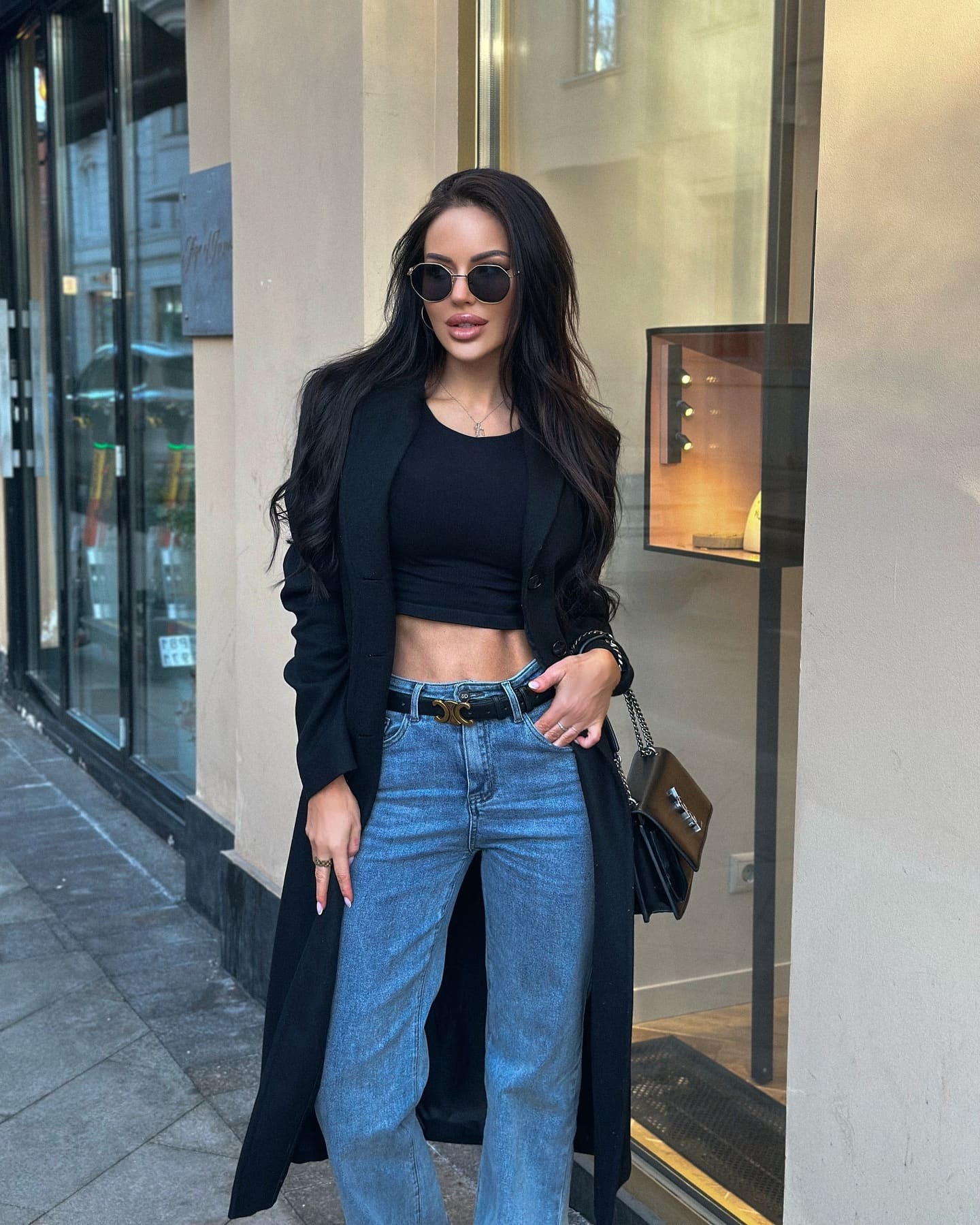 Marika stands on a city sidewalk in a black cropped top, long coat, and jeans, holding a handbag in front of a storefront.