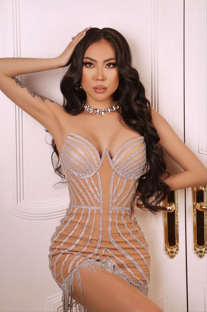 Doramia wears a nude rhinestone mini dress and choker, posing with a hand on her head before a white paneled door.