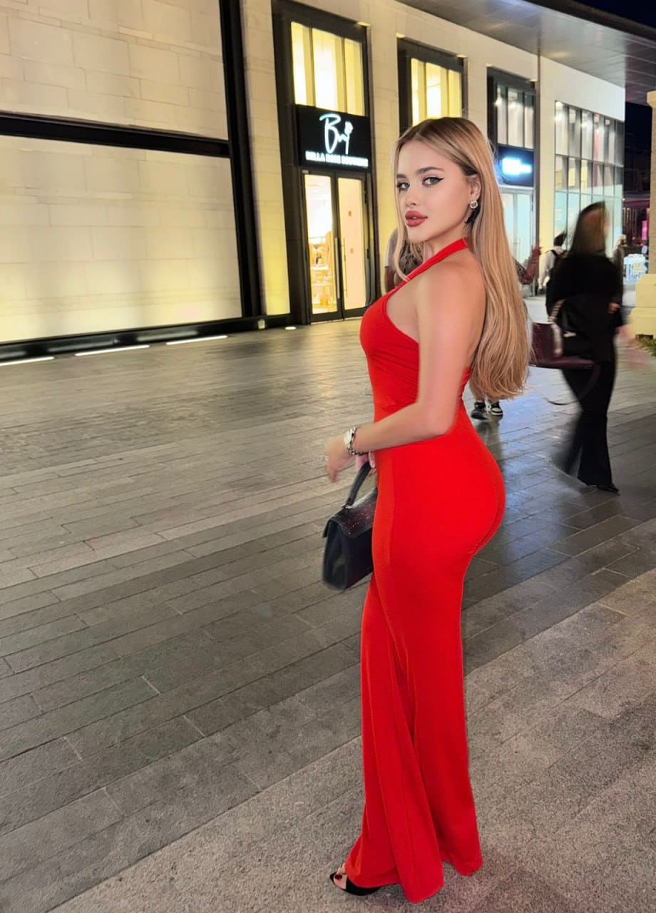A woman stands in profile on a night walkway wearing a red halter maxi dress and holding a black handbag near storefronts.