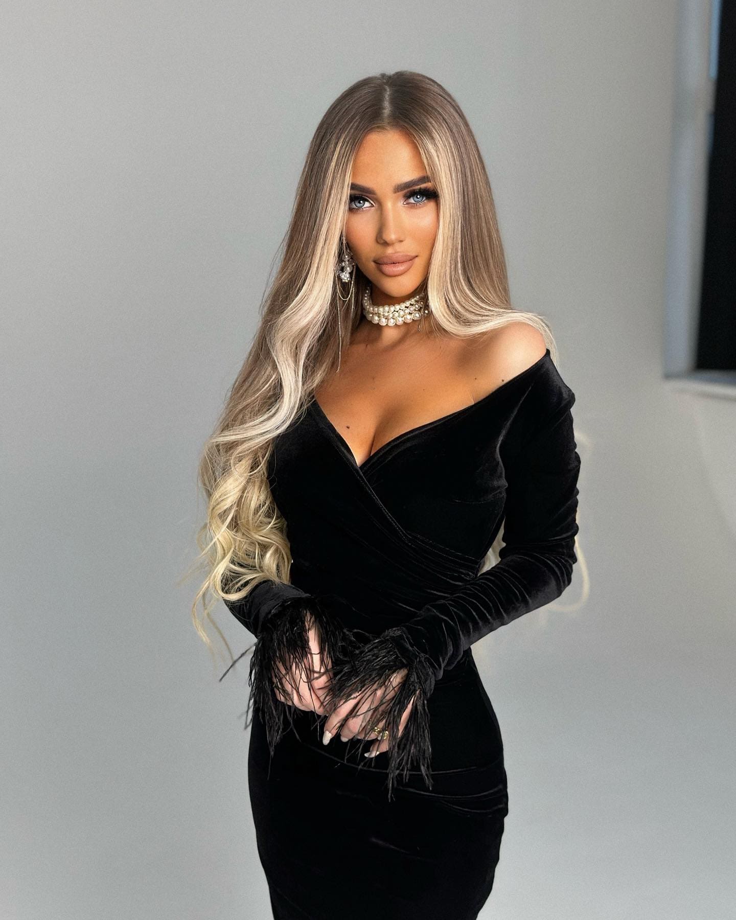 Sonna stands in a black velvet off-shoulder dress with fringed cuffs and a pearl choker against a gray studio background.
