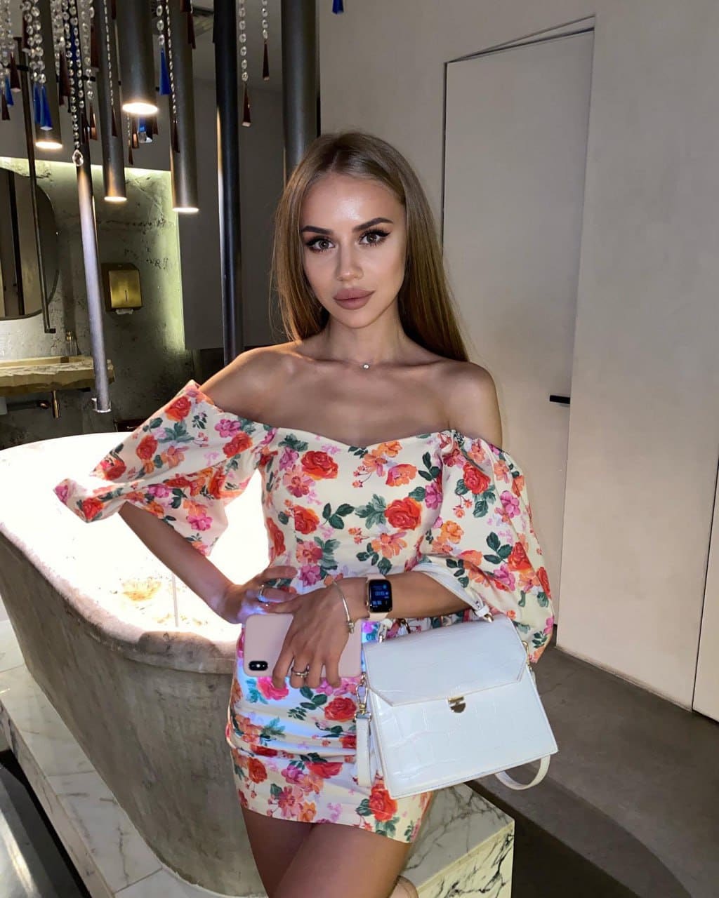 A woman with brown hair leans against a stone bathtub indoors, wearing an off-the-shoulder floral dress and holding a white handbag.