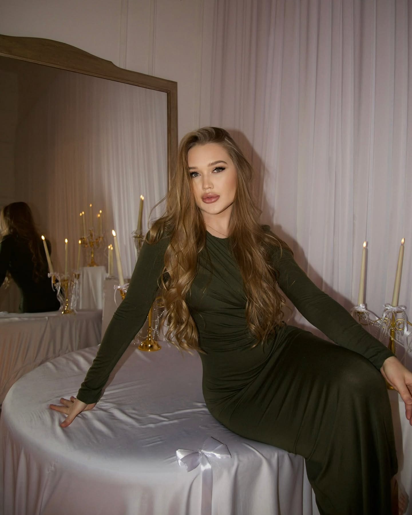 Viona sits on a white-draped table in a dark green dress, with candles and a mirror behind her.