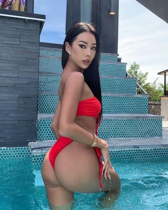 A woman in a red bikini stands in a pool with her back partially turned, near tiled steps and a stone wall.