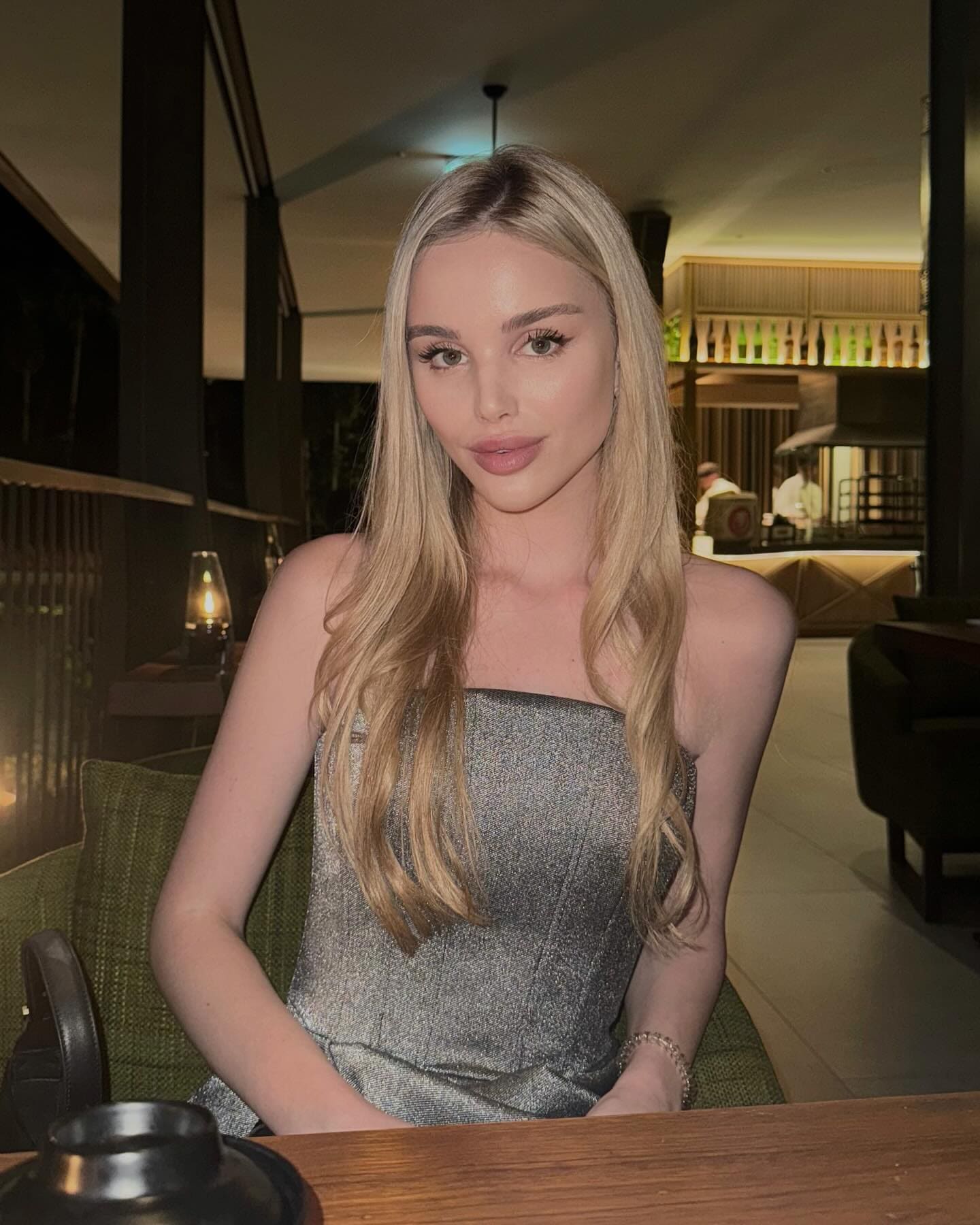 Marilli sits at a wooden table in a dim restaurant, wearing a strapless silver-grey dress with arms resting on the surface.