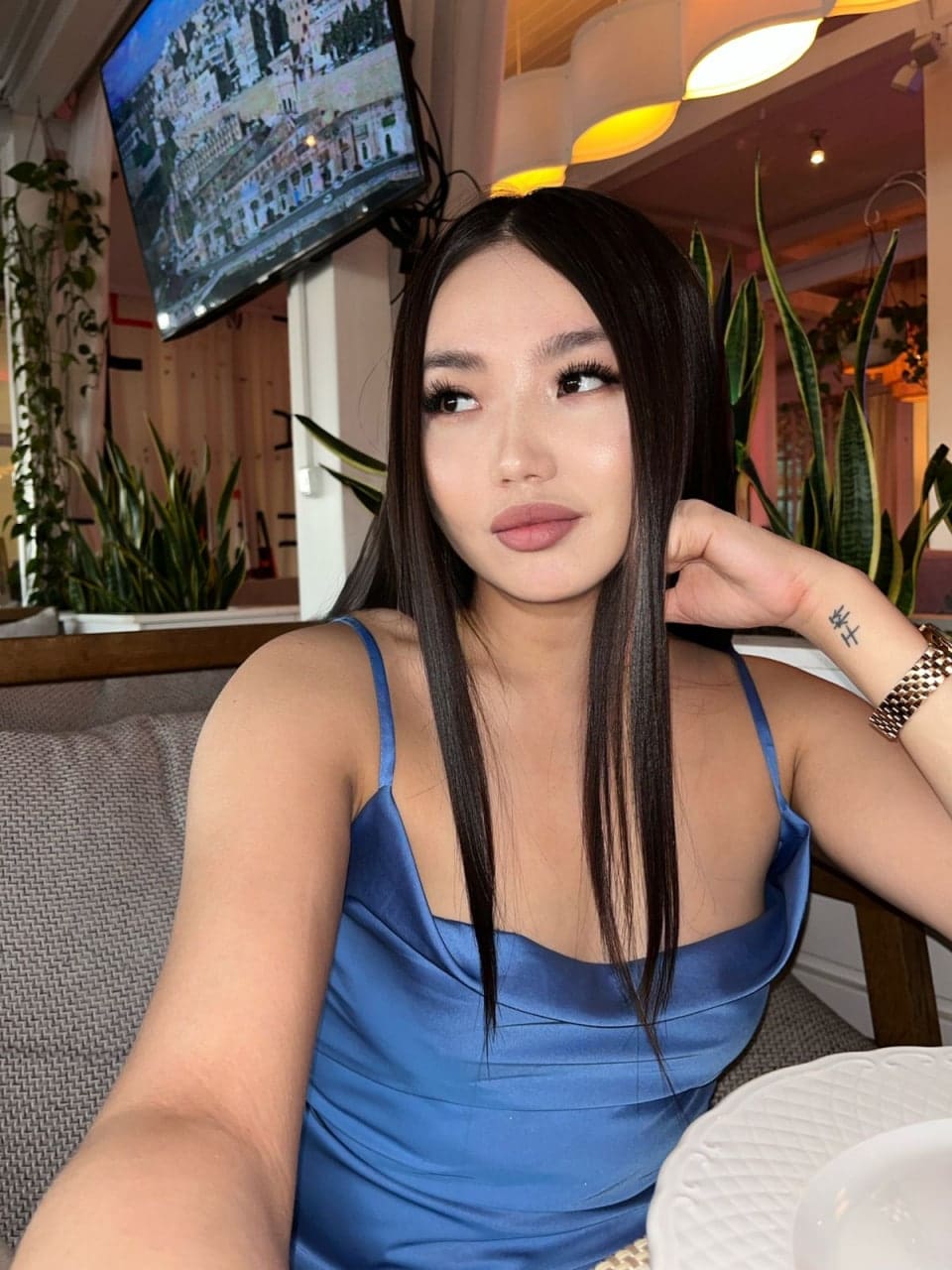 Karina sits on a couch in a blue satin slip dress, hand near her face, with plants and a TV in the background.