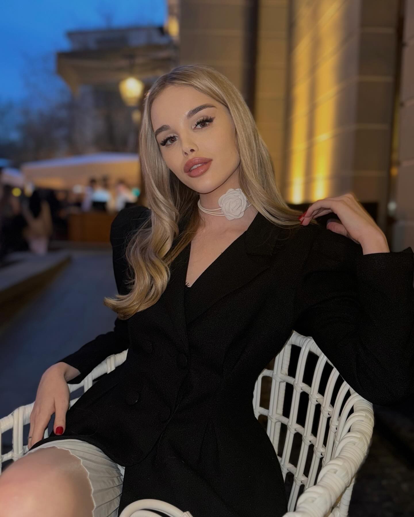 A woman in a black blazer and light skirt sits in a white wicker chair outdoors at night, wearing a white flower choker.