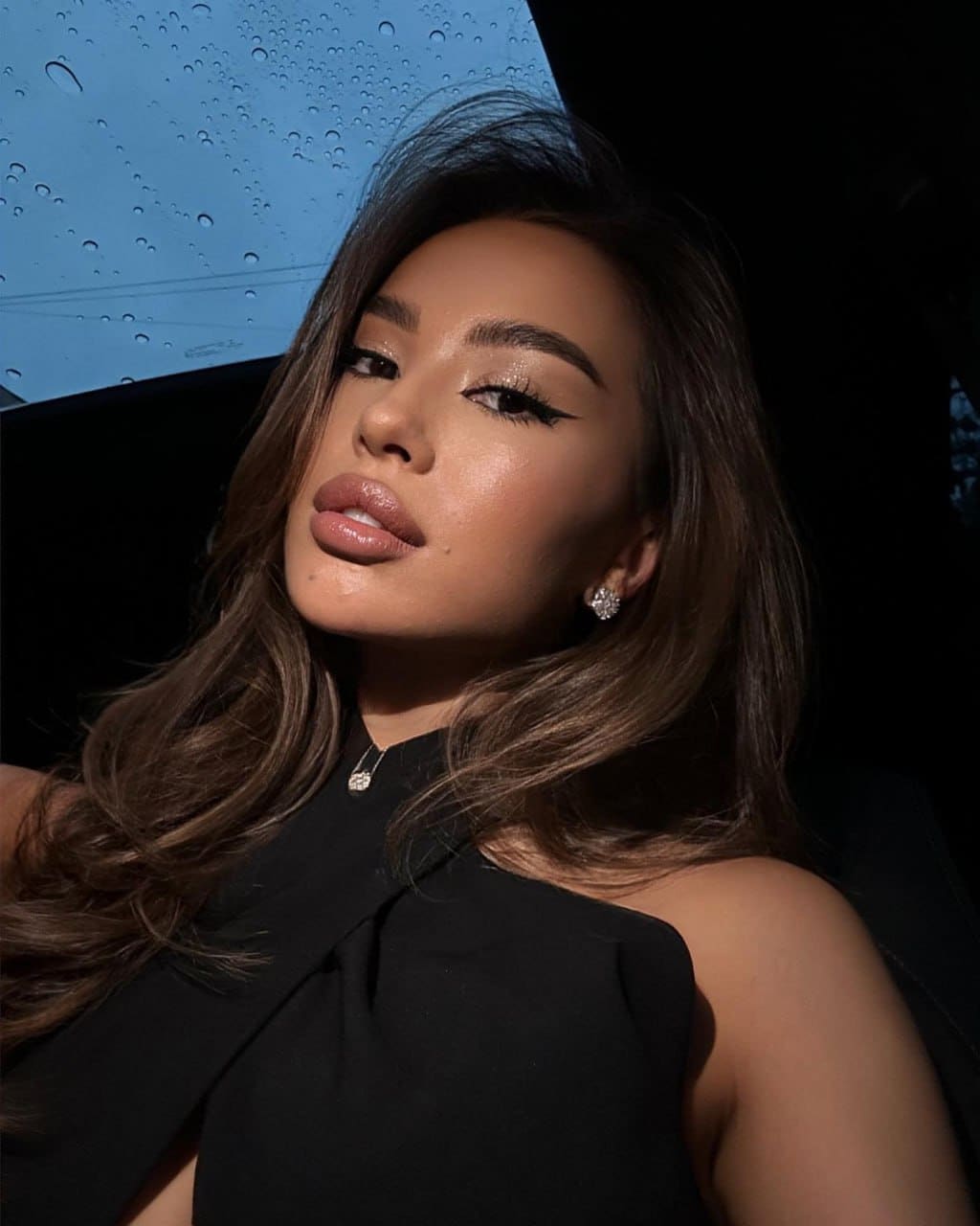 Vasilina sits in a car wearing a black sleeveless top and pendant necklace, with a rain-streaked window behind her.